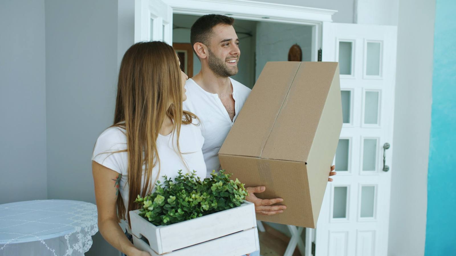 A smiling couple carries a moving box and a plant into their new home